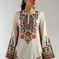 Woman wearing a cream-colored embroidered dress with colorful patterns on a plain background